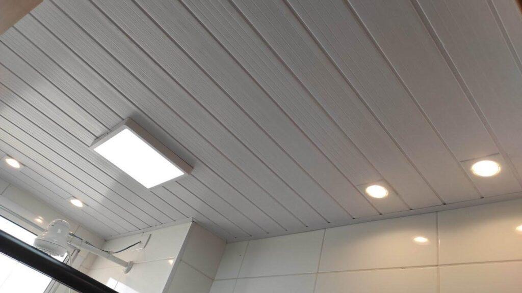 Teto de banheiro com forro de PVC branco da PERMATTI. O acabamento em réguas possui iluminação embutida com plafon e spots, sobre parede de azulejos brancos.