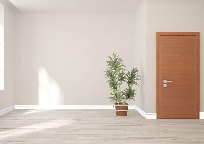 Ambiente interno clean com piso amadeirado claro e paredes em cinza suave. Um rodapé de PVC Permatti branco faz o acabamento na base da parede, conferindo um design moderno e sofisticado. Uma planta em vaso e uma porta de madeira clara completam o cenário.